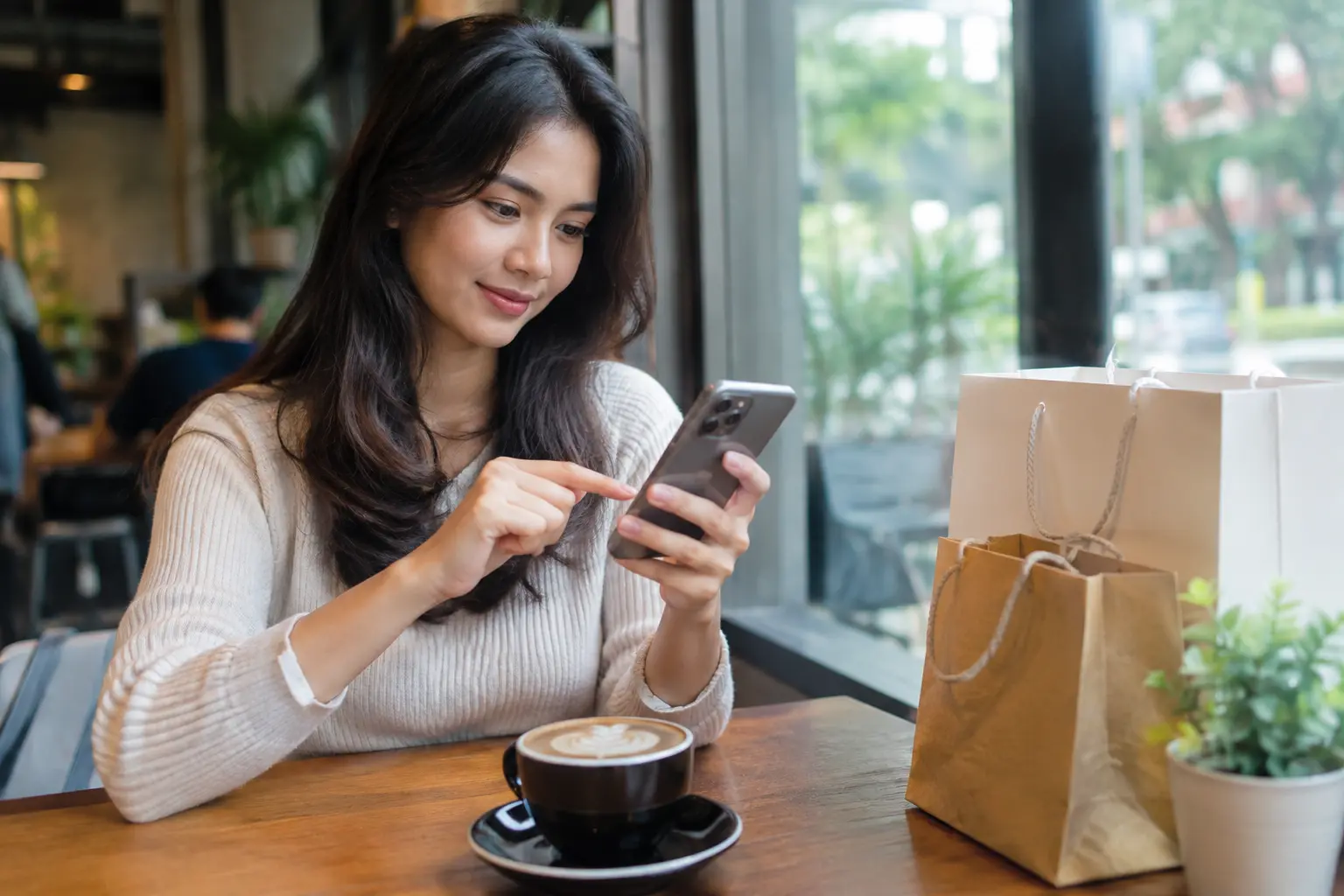 Perempuan muda menelusuri toko online lewat ponsel di meja kafe dengan kantong belanja